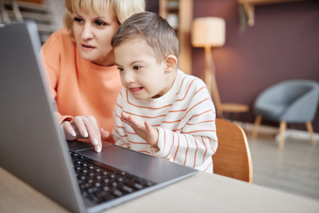 Closeup of excited little boy with down syndrome using computer at home mother or tutor assisting