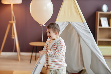 Portrait of cute little boy with down syndrome holding balloon while playing at home and laughing, copy spaceの写真素材