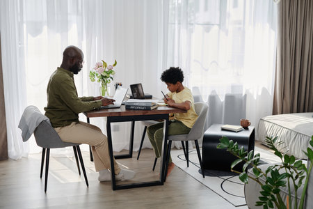 Child doing his homework and his dad working online on laptop while they sitting at table in the...