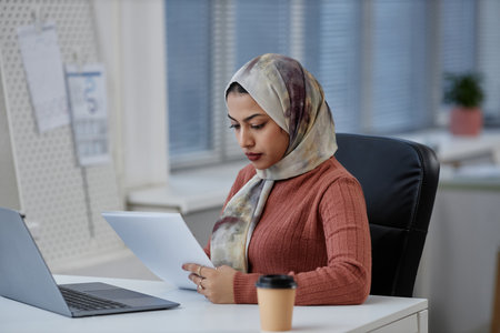 Young serious Muslim female entrepreneur or student in headscarf looking through document while sitting by workplace in front of laptopの写真素材