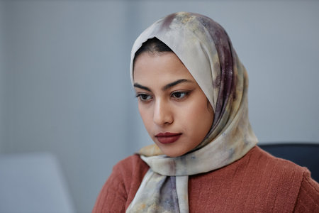 Young pretty Muslim female employee in headscarf looking at laptop or other gadget screen while...