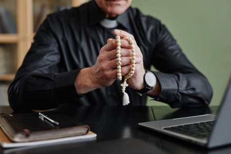 Closeup of senior priest holding rosary while praying at desk in office, copy spaceの写真素材
