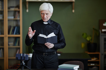 Waist up portrait of white haired senior priest holding Bible and recording video for online blog in...