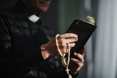 Side view closeup of unrecognizable priest holding Bible and rosary while praying in dramatic light, copy spaceの写真素材