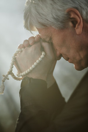 Vertical closeup portrait of white haired senior priest praying with rosary and serene lightingの写真素材