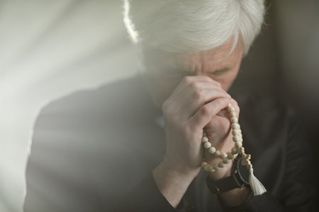 Front view of white haired senior priest praying with rosary in serene god rays, copy spaceの写真素材
