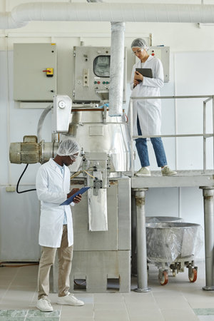 Vertical wide angle view at two people wearing lab coats standing in factory workshop on different...