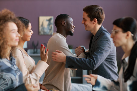 Two happy intercultural guys shaking hands and giving each other hug at home party while standing...