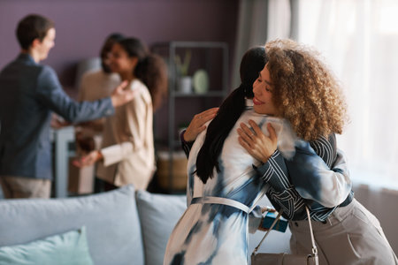 Focus on two young cheerful girls in casualwear giving each other hug while standing in front of camera against group of intercultural peopleの写真素材