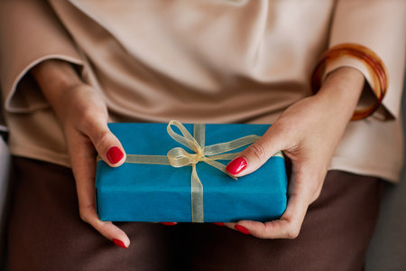 Hands of young woman in beige silk blouse holding small blue giftbox with birthday present while sitting in front of camera at partyの写真素材