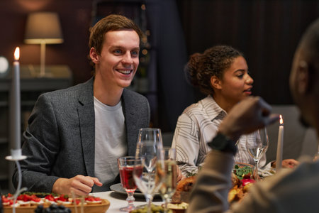 Happy young man looking at his buddy during conversation by festive dinner while sitting in front of him by served table at home partyの写真素材