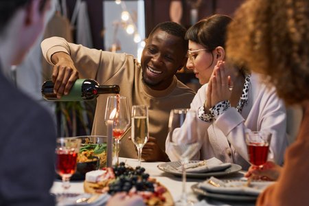 Happy young African American man pouring red wine into flute of his girlfriend while sitting next to...