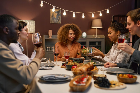 Group of young intercultural men and women sitting by served festive table, communicating and having homemade food and drinksの写真素材
