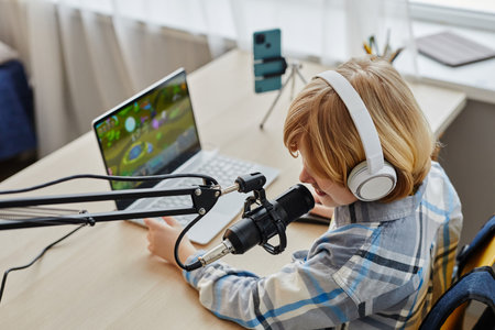 Blond little boy in headphones speaking in microphone while sitting by desk in front of laptop with...