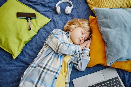 Above angle of tired blond schoolboy and blogger napping on his bed among group of pillows, laptop, smartphone and headphonesの写真素材