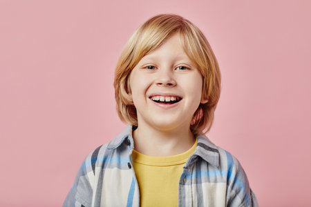 Cute laughing blond boy in checkered shirt and yellow t-shirt looking at camera with toothy smile while standing on pink backgroundの写真素材