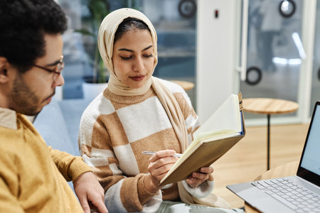 Portrait of young Muslim woman wearing head scarf in office and talking to male colleague discussing notesの写真素材