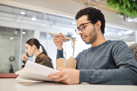 Side view portrait of young Middle Eastern man eating takeout food at office workplace and reading documents, copy spaceの写真素材