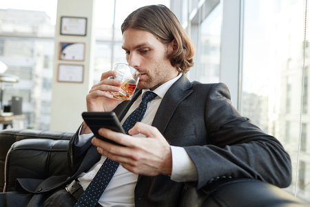 Portrait of handsome businessman drinking whiskey in office and checking text messagesの写真素材