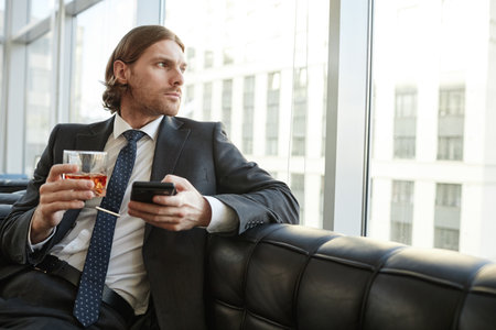 Portrait of handsome businessman drinking whiskey in office and looking at window relaxing on leather couch, copy spaceの写真素材