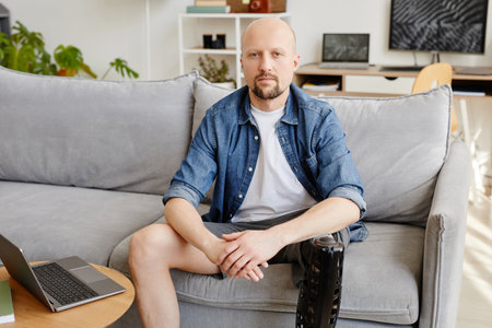 Front view portrait of adult man with prosthetic leg looking at camera while sitting on sofa at...