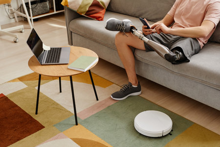 Closeup of man with prosthetic leg using smartphone while relaxing on couch at home robot vacuum on floor, copy spaceの写真素材