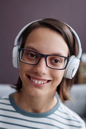 Vertical closeup portrait of smiling teen girl with disability wearing headphones at home and looking at cameraの写真素材