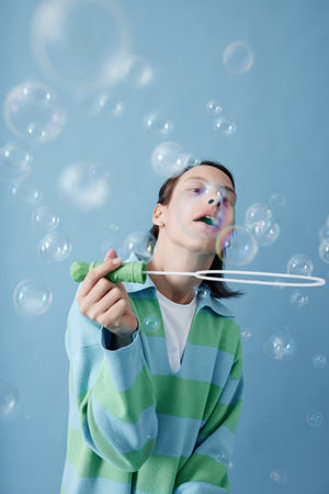 Waist up portrait of smiling teenage girl with disability blowing bubbles against blue background