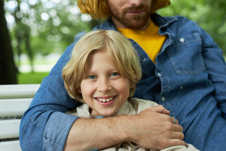 Closeup of father and son sitting on bench in park together with blonde boy smiling at camera
