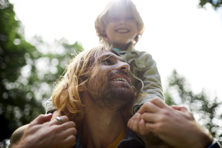 Closeup portrait of happy young father carrying son on shoulders outdoors in sunlightの写真素材