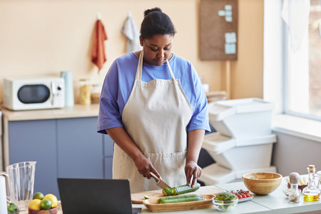 Waist up portrait of adult black woman wearing apron in kitchen and cutting vegetables while following cooking video online, copy spaceの写真素材