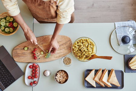 Top view background image of woman cutting cherry tomatoes and broccoli on wooden cutting board in kitchen while cooking pasta, copy spaceの写真素材