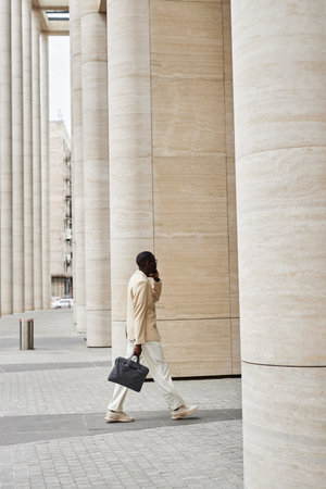 Side view of young elegant male entrepreneur in elegant white suit talking on mobile phone while moving towards huge modern buildingの写真素材