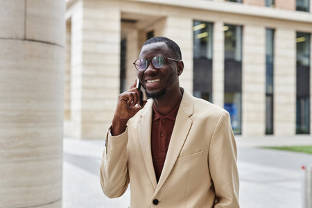 Young successful African American businessman in ivory jacket speaking on smartphone while standing in front of camera in urban environmentの写真素材