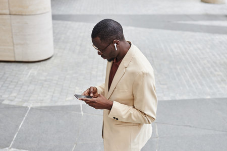 High angle of young black businessman in earphones and pastel color suit texting in mobile phone or scrolling through playlist in the streetの写真素材