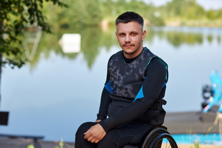Portrait of young man with disability looking at camera sitting in wheelchair by lake and wearing...