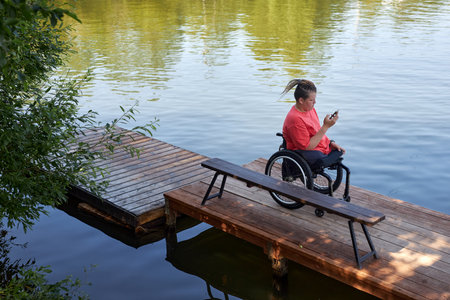 High angle portrait of young man with disability sitting in wheelchair by lake and using smartphone,...