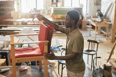 Side view portrait of young black man as craftsman refurbishing old chair in carpentry workshop, copy spaceの写真素材