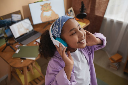 High angle portrait of smiling young black girl wearing headphones at home and listening to music...
