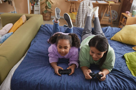 High angle portrait of happy black mother and daughter playing video games together lying on bed at...