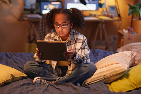 Full length portrait of teen black girl with pigtails using digital tablet while sitting on bed at...