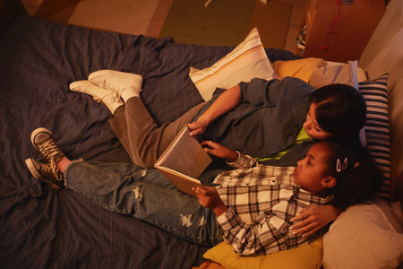 Portrait of young African American mother and daughter lying in bed together and reading book at nightの写真素材