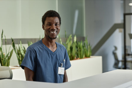 Happy young male clinician or assistant in blue medical uniform standing by reception counter in front of camera and meeting new patientsの写真素材
