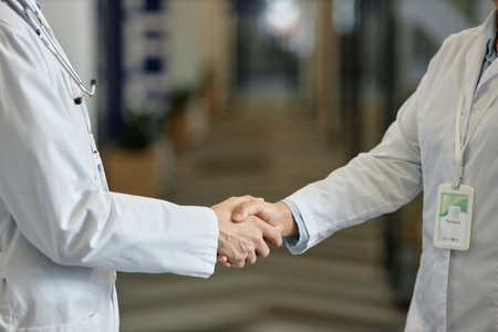 Close-up of hands of two general practitioners in lab coats standing in front of camera in corridor of modern hospital during handshakeの写真素材
