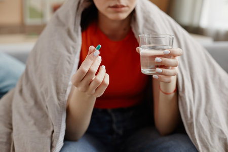 Closeup of young woman holding capsule pill and glass of water wrapped in blanket while taking...