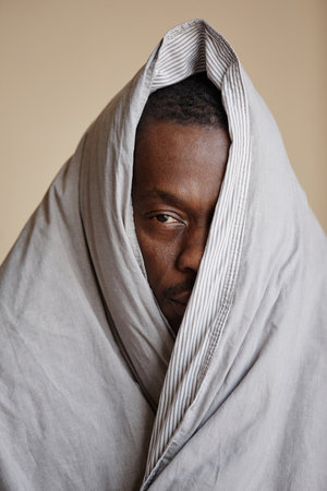 Minimal portrait of depressed Black man wrapped in blanket looking at camera struggling with mental health issuesの写真素材