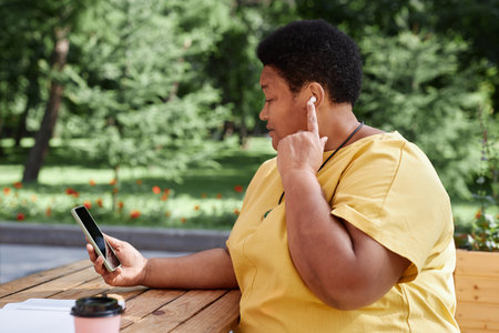 Side view portrait of modern African American senior woman using smartphone outdoors and tapping wireless earphones, copy spaceの写真素材