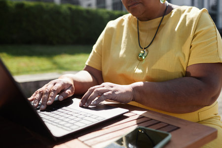 Closeup of mature Black woman using laptop outdoors in sunlight focus on hands typing, copy spaceの写真素材