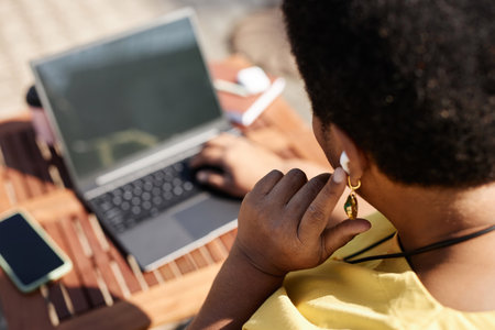 Closeup of Black woman tapping wireless earphones while working with laptop outdoors, copy spaceの写真素材