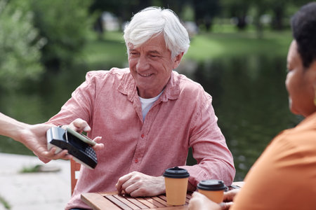 Portrait of smiling senior couple paying via NFC by smartphone at outdoor cafe, copy space